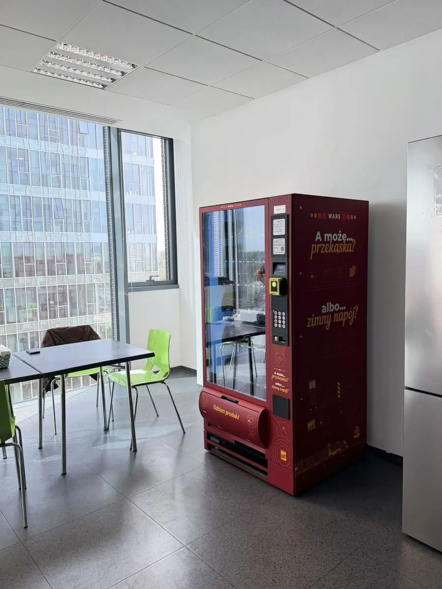 Bordowy automat vendingowy obok stolików i krzeseł w jasnym pomieszczeniu